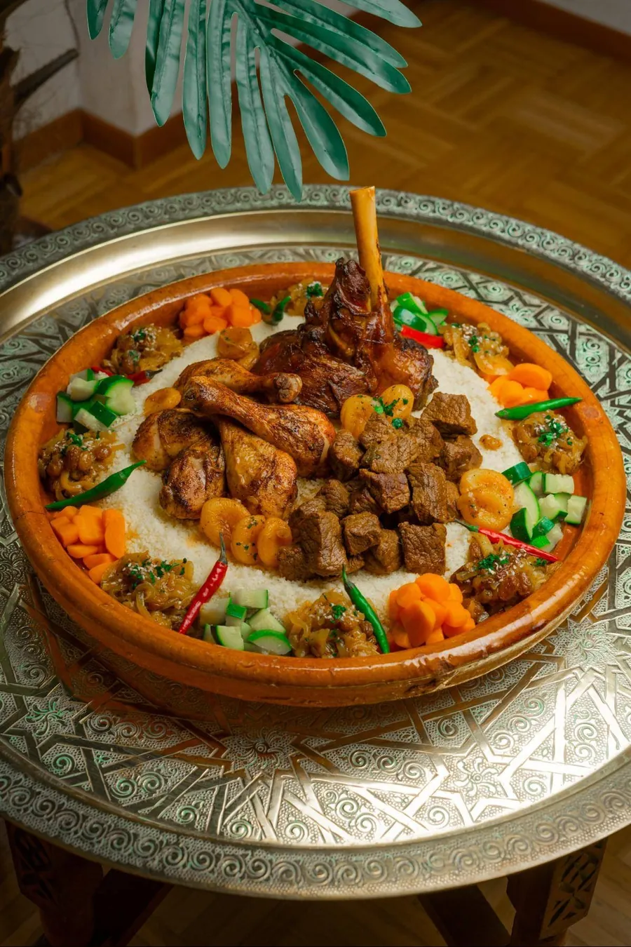 Grand plat de couscous royal posé sur un plateau en cuivre ciselé, sous une feuille de palmier.